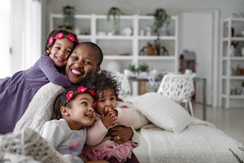 A smiling person embraces three young children in a bright, modern living room.