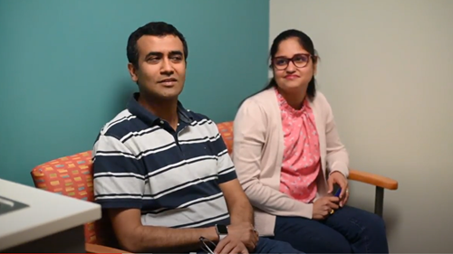 A person in a striped polo shirt and another in a pink top and light cardigan sit on a sofa in a room with teal walls.