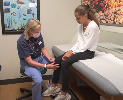 A healthcare provider in a face mask performs a knee examination on a patient sitting on an exam table in a clinical room.
