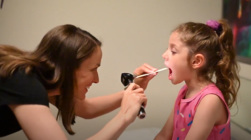 A healthcare provider examines a young patient’s throat using a tongue depressor and an otoscope.
