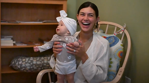 A person sitting in a rocking chair holding a baby wearing a white headwrap in front of a wooden bookshelf.