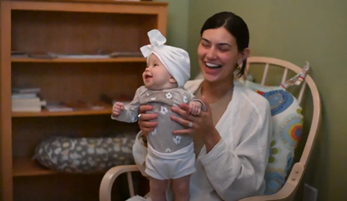 A person holds a smiling baby in a white hat while sitting in a wooden rocking chair in a room with a bookshelf.
