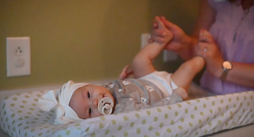 A person holds an infant’s feet while changing them on a patterned changing pad in a room with a wall outlet.