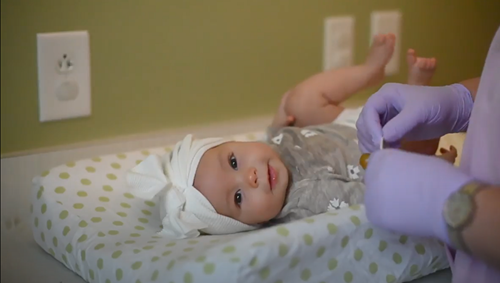 An infant wearing a white headband lies on a polka-dot pad while a person in purple gloves prepares a medical procedure.