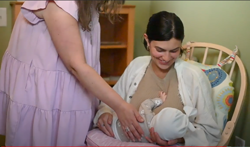 A person in a rocking chair cradles a baby while another person in a light purple dress gently touches the baby.