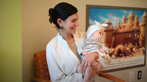 A person smiling while holding a baby wearing a white hat and pacifier, sitting in a chair near a wall art of a castle.