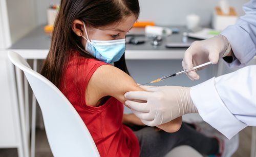 A healthcare professional wearing gloves administers an injection into the upper arm of a person wearing a medical mask.