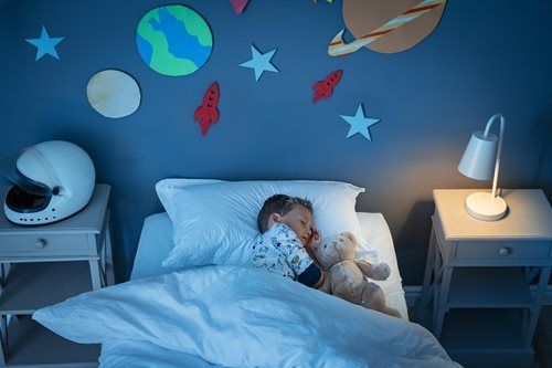 A child sleeps in a bed under space-themed wall decorations, with a bedside lamp on and a white helmet on a nightstand.