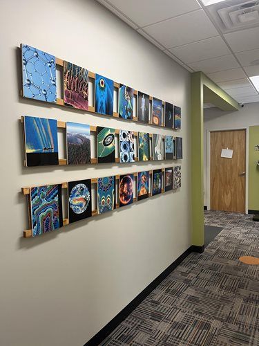 Three rows of framed, colorful abstract and scenic art prints mounted on a light-colored office hallway wall.