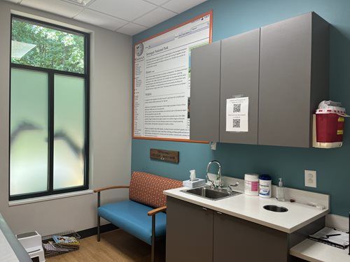 A clinical examination room with a blue bench, a small sink, wall cabinets, and a window with frosted glass.