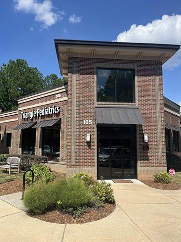 The brick exterior of Triangle Pediatrics, unit 105, featuring large windows, a metal awning, and a sunny blue sky.