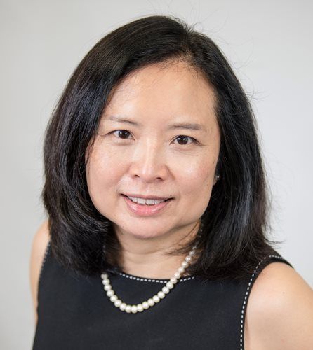 Headshot of a smiling person with dark hair, wearing a pearl necklace and a black sleeveless dress against a grey wall.