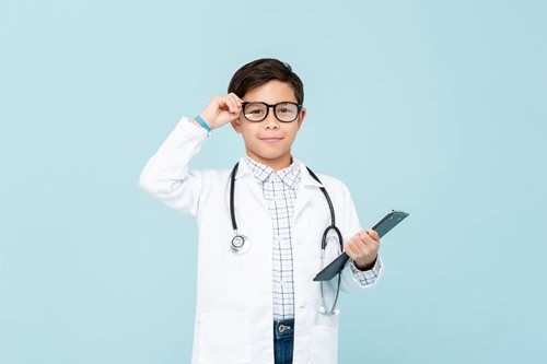 A child dressed as a doctor in a white lab coat with a stethoscope around their neck, adjusting glasses and holding a pad.