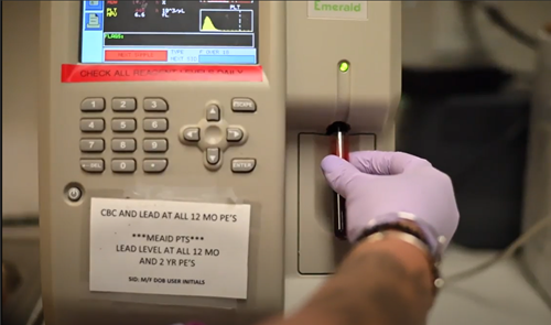 A gloved hand inserts a test tube into an automated blood analyzer labeled with instructions for pediatric lead testing.