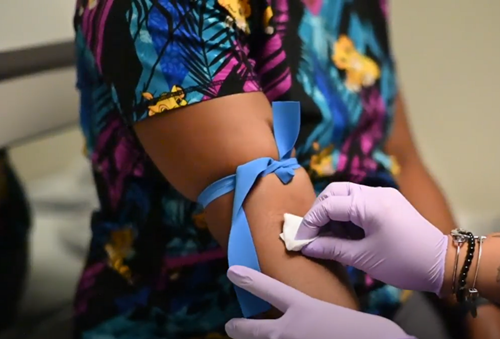 A healthcare worker wearing purple gloves cleans a patient's arm with an alcohol swab before a blood draw.