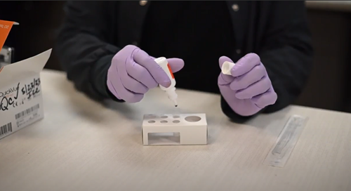 A person wearing purple medical gloves uses a dropper to place liquid into a small testing device on a desk.