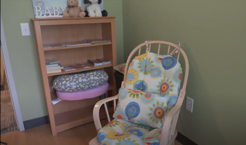 A light wood rocking chair with a floral cushion sits next to a wooden bookshelf with toys and books against a green wall.