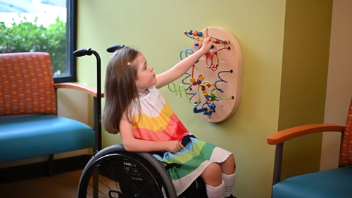 A child in a wheelchair plays with a wall-mounted bead maze in a room with orange chairs and a green wall.