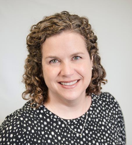 A professional headshot of a smiling woman with curly hair wearing a black-and-white patterned top.