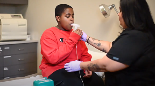 A healthcare provider in black scrubs assists a patient in a red sweatshirt using a nebulizer mask in a clinic.