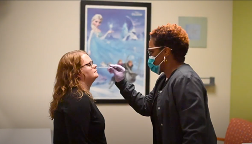 A healthcare worker in a face mask and gloves administers a nasal swab test to a person in an office with Frozen decor.