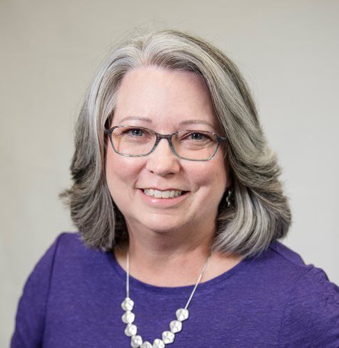 Smiling person with gray hair and glasses, wearing a purple top and a silver beaded necklace against a neutral background.