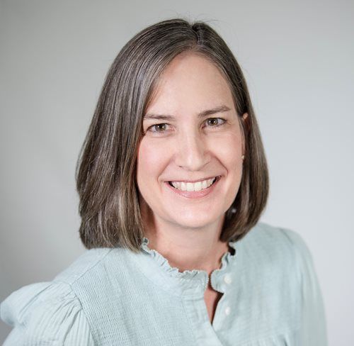 A person with shoulder-length gray-brown hair smiles warmly, wearing a light blue collared blouse against a gray background.