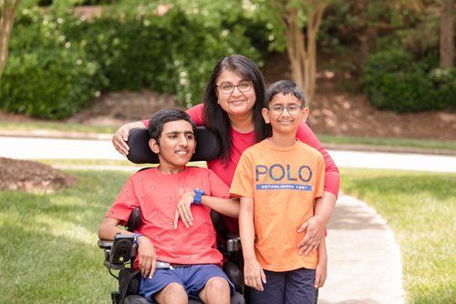 A person in a wheelchair and another person stand beside a woman outdoors, all smiling in a green park setting.