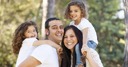 A happy family of four smiling outdoors, with two children being carried on the shoulders of the two adults.