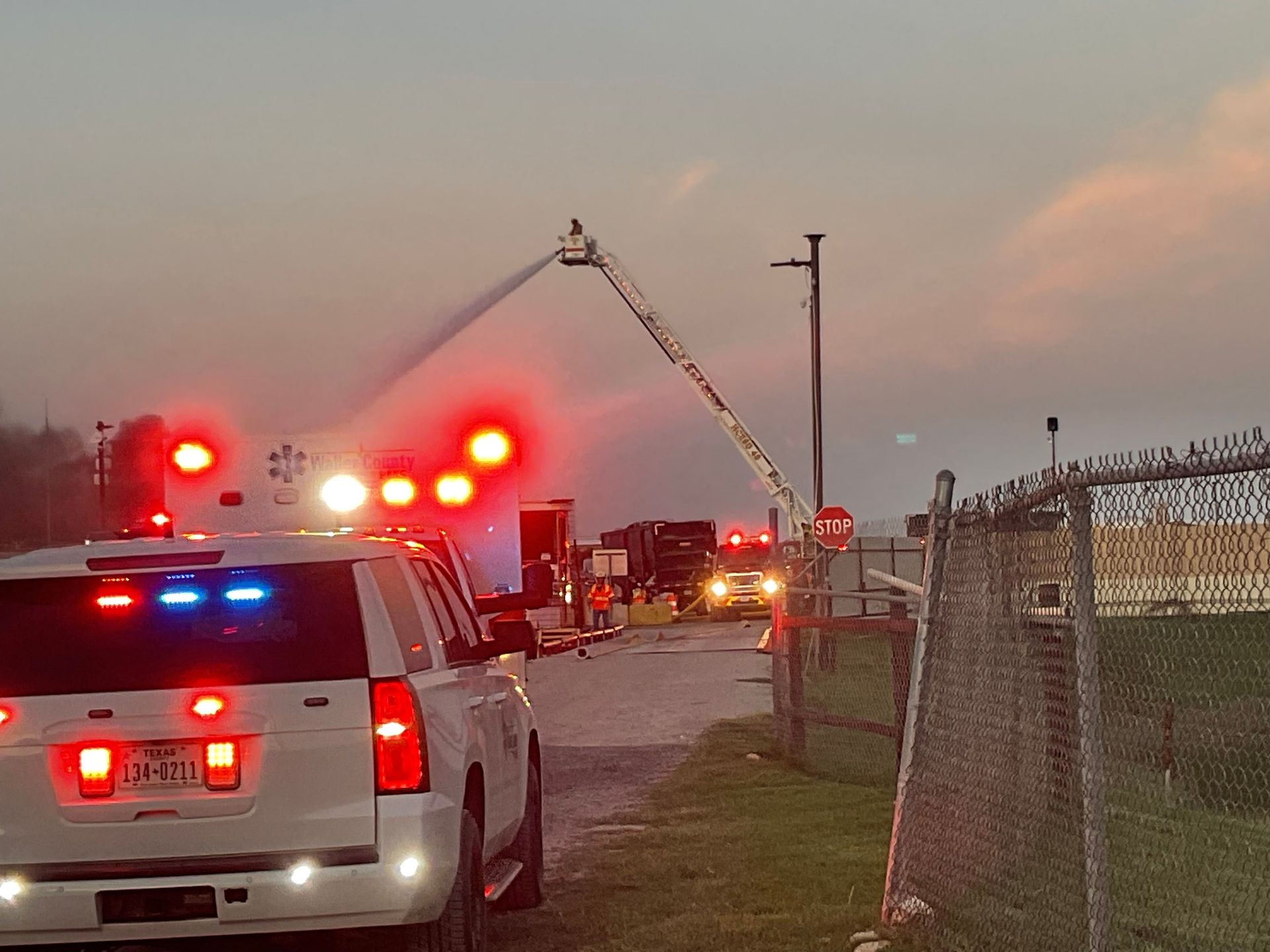 A fire command vehicle is parked on a road with an aerial fire truck with a ladder spraying water.