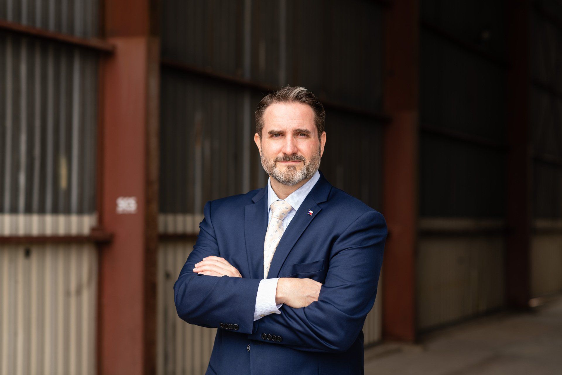 A man in a suit and tie is standing with his arms crossed in front of a building.