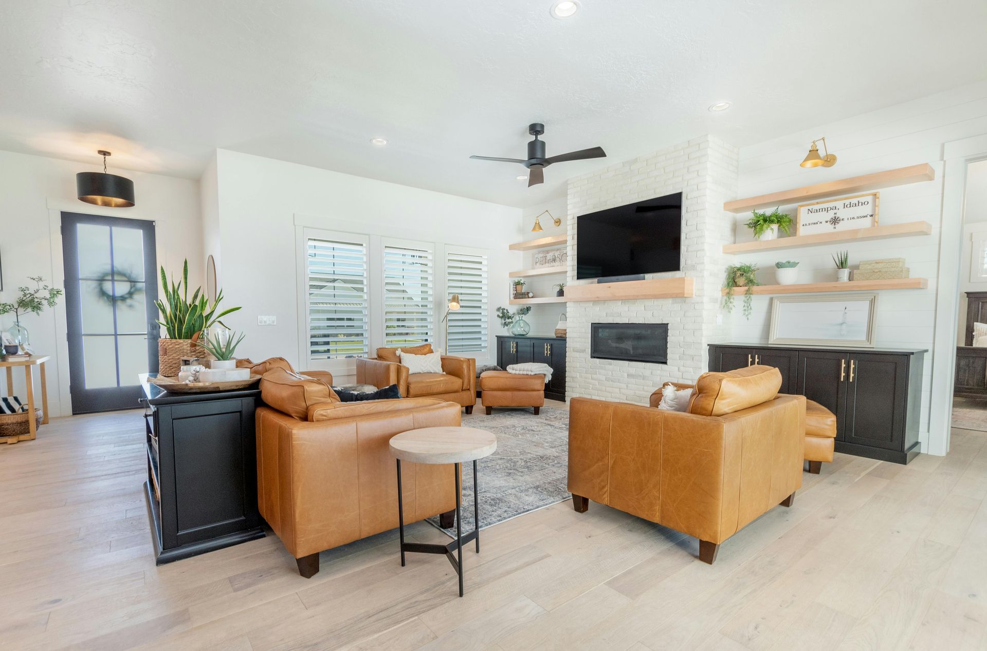 A bright living room with two tan leather chairs, a stone fireplace, floating shelves, and a dark wood cabinet.