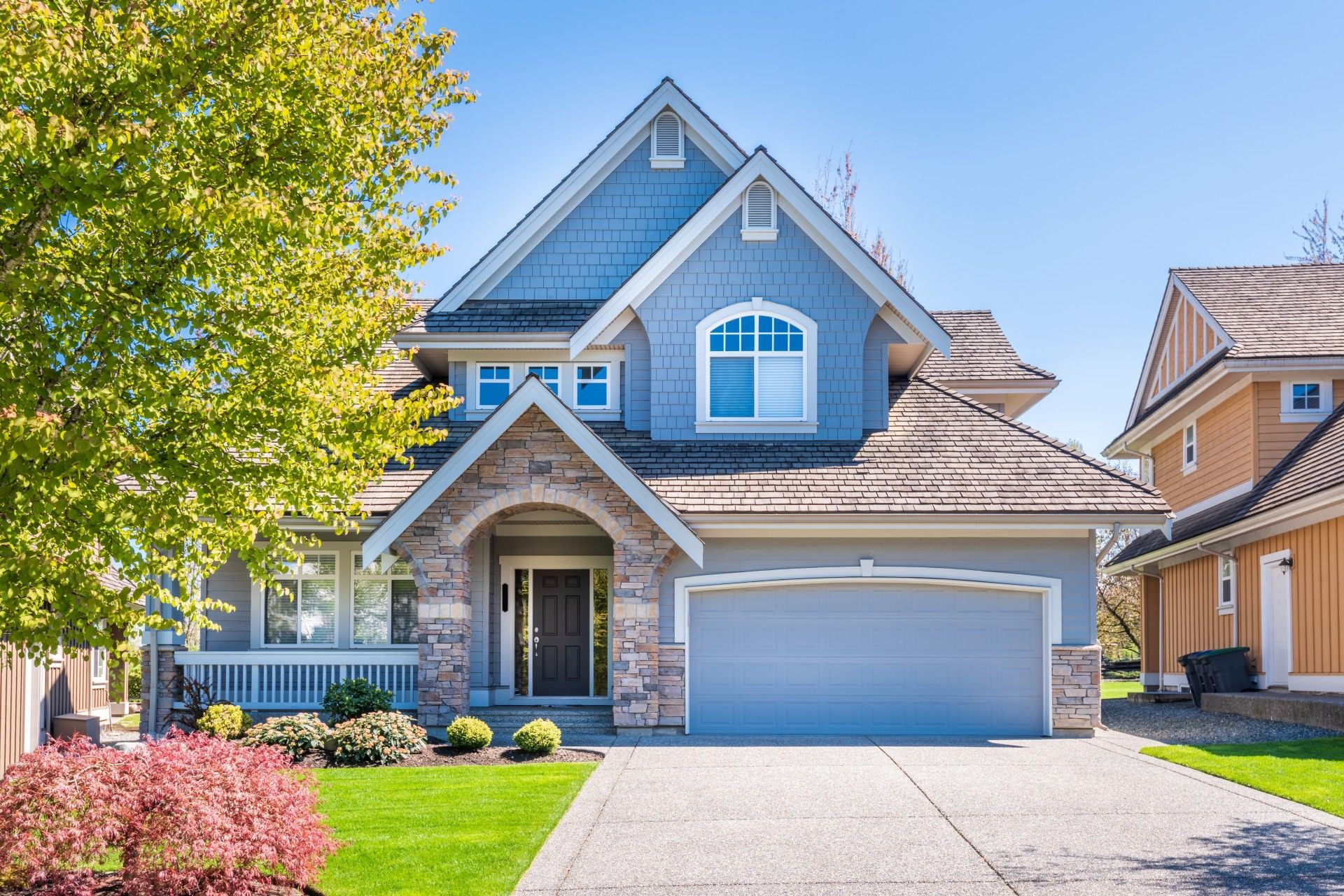A two-story blue house with stone accents, a garage, a front porch, and a driveway on a sunny day.