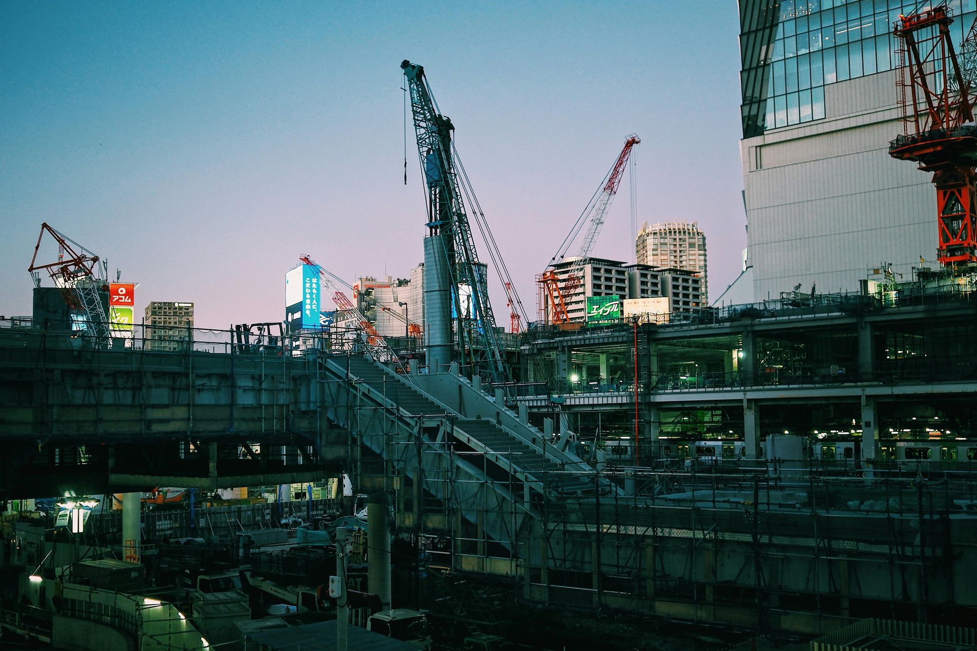 Dusk cityscape with construction cranes, elevated walkways, and buildings under a blue sky