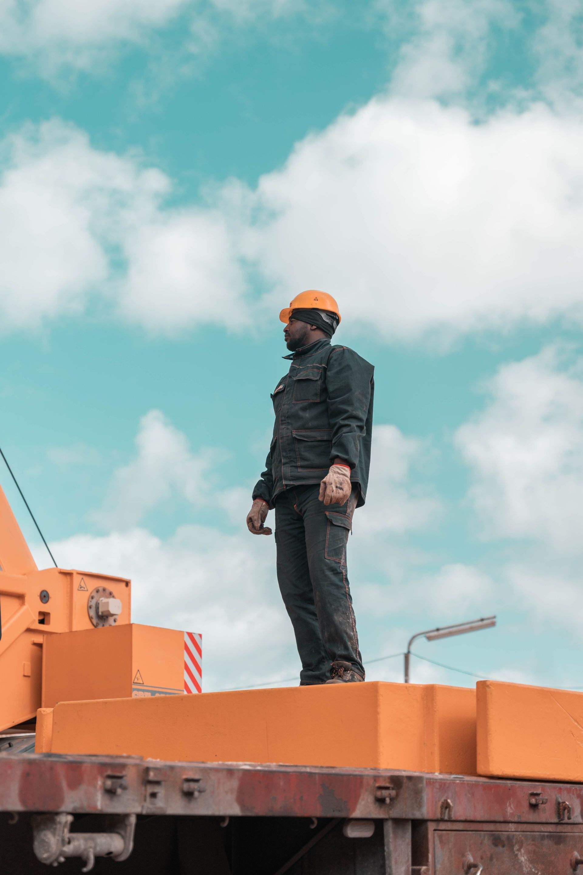 Worker in orange hard hat standing on orange machinery against a cloudy sky