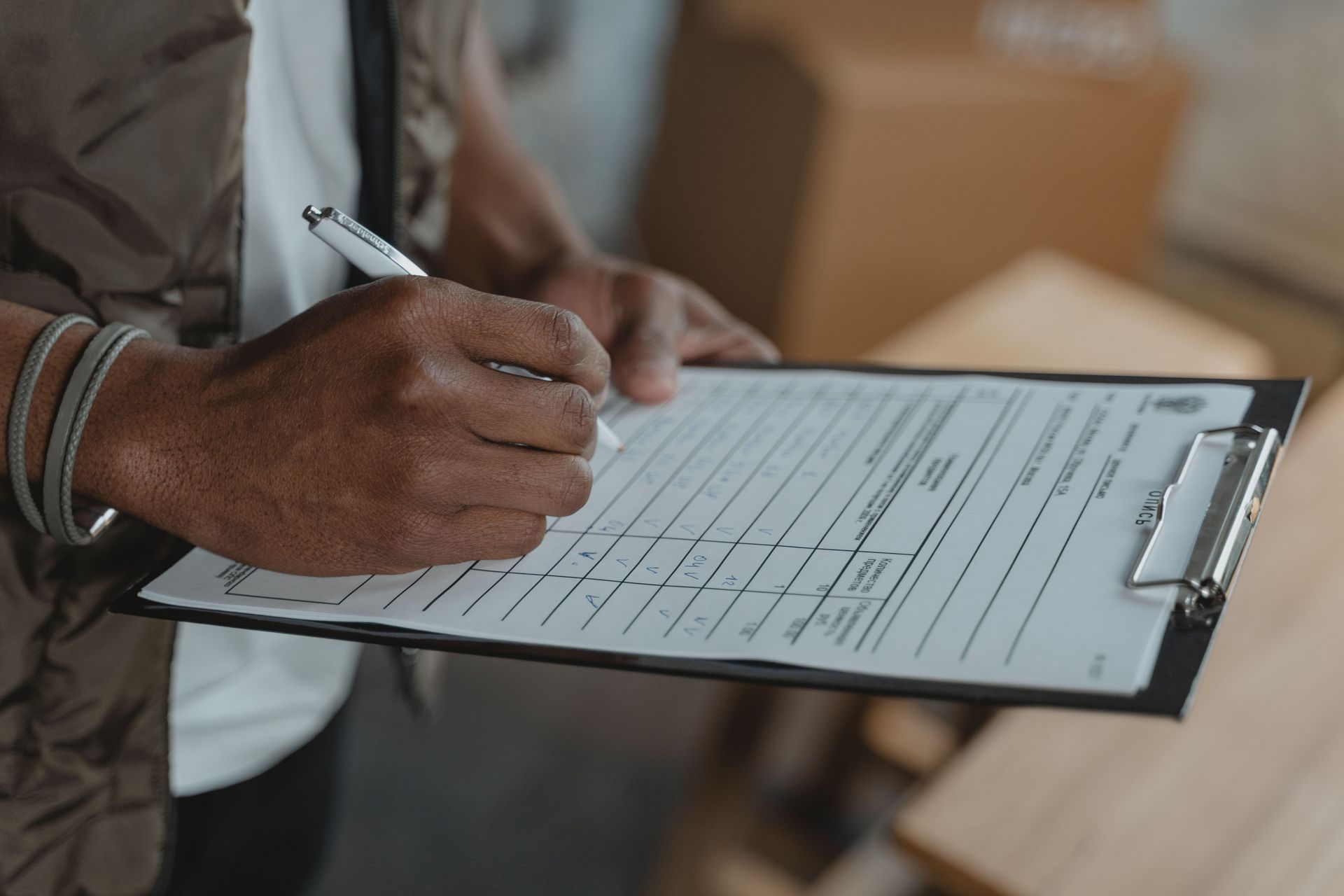 Person writing on a clipboard form indoors, with a pen and a table in the background.