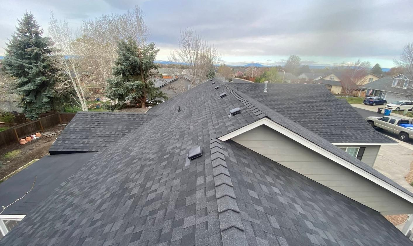 Dark gray asphalt shingle roof on a house, with trees and cloudy sky in the background.