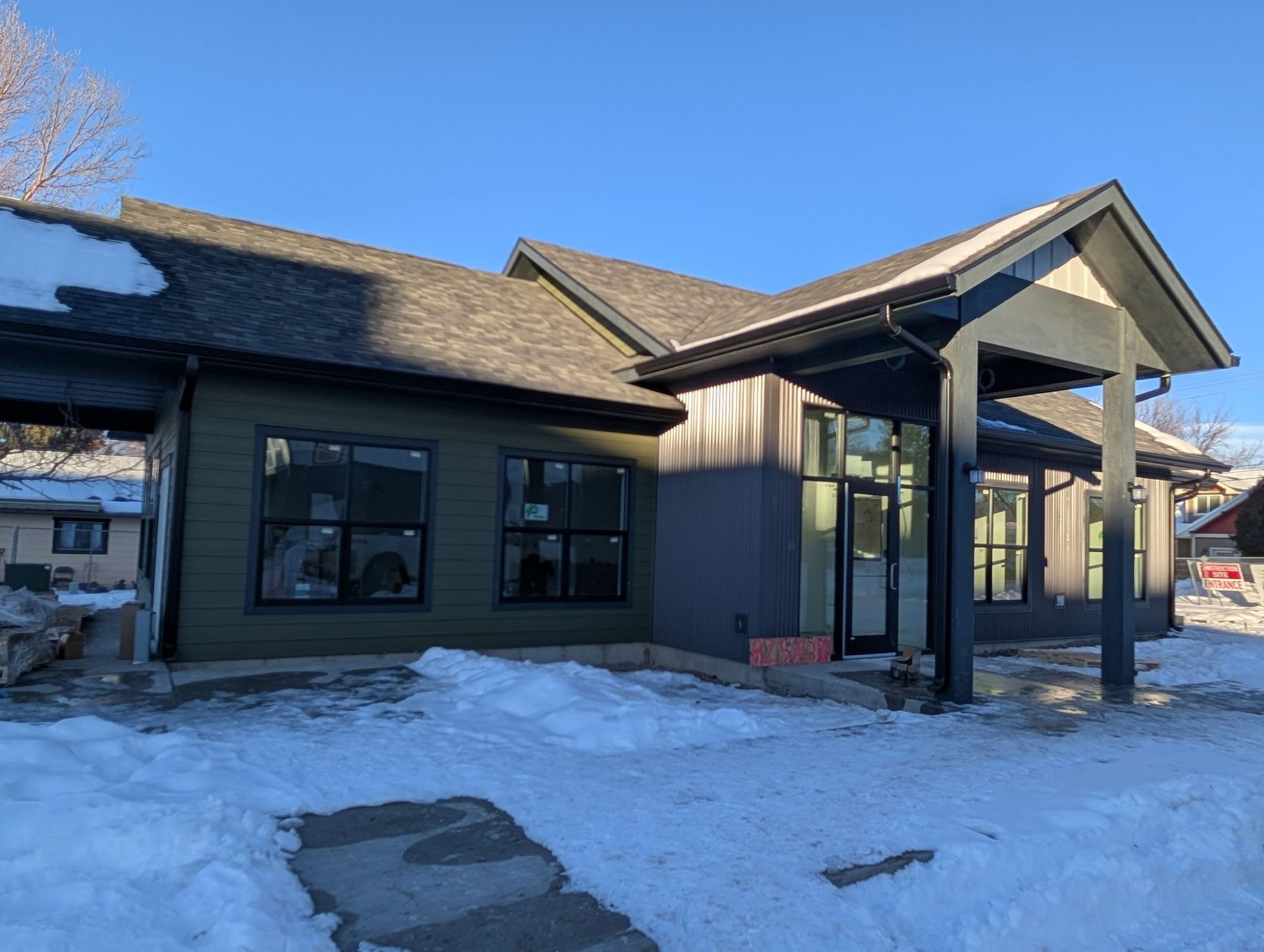 A modern building with green and gray siding, black window frames, and snow in the foreground.