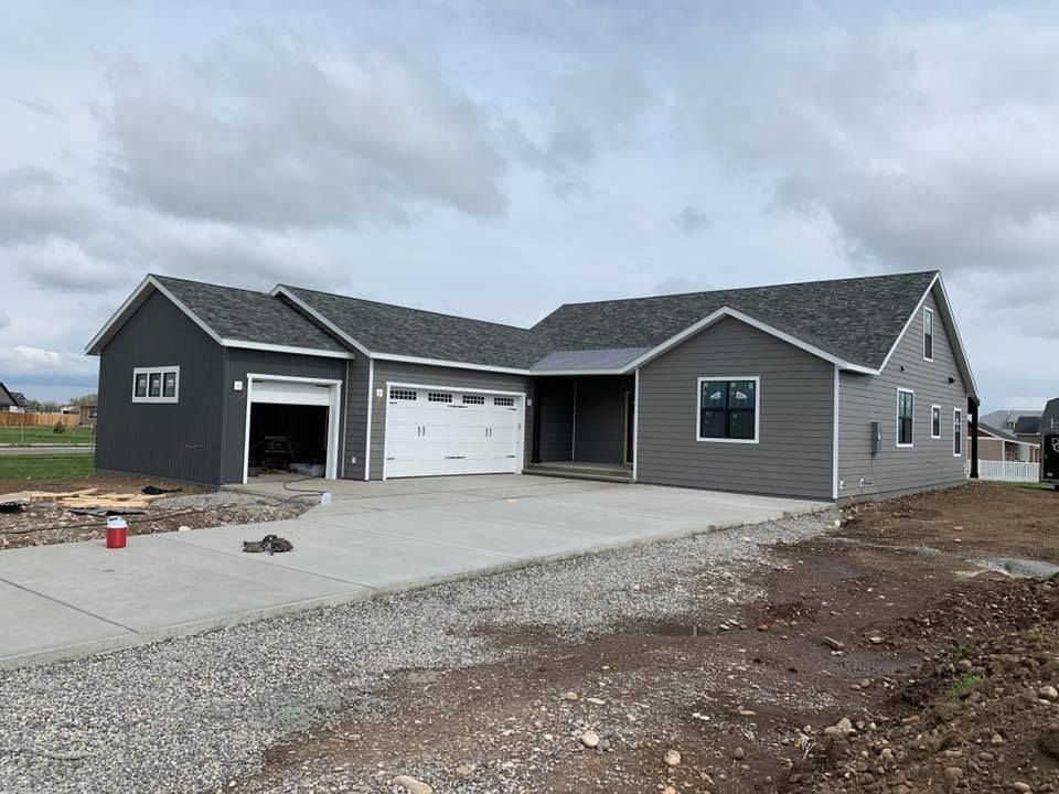 Gray house under construction, with open garage door and concrete driveway. Cloudy sky overhead.