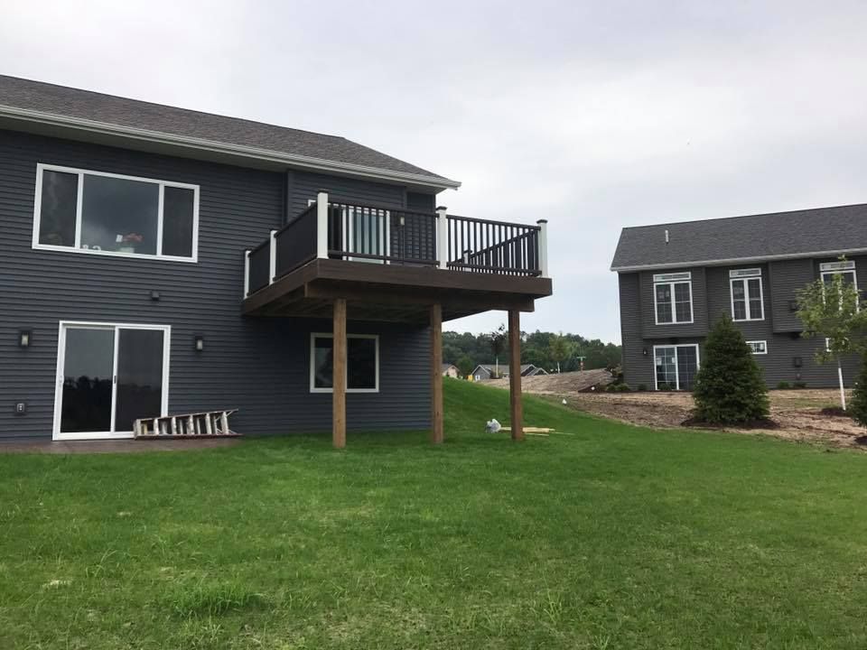 Two-story house with dark siding and a wooden deck, located in a grassy yard. A second house is in the background.