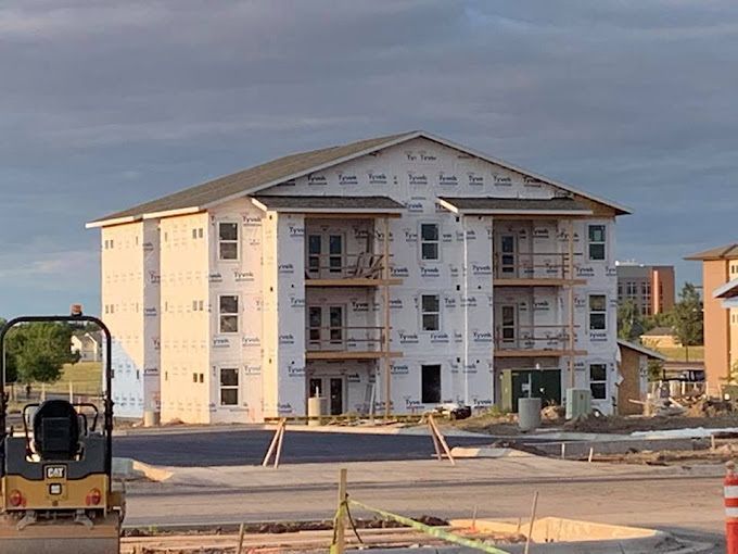 Apartment building under construction, wrapped in white material, with balconies. Road and equipment in foreground.
