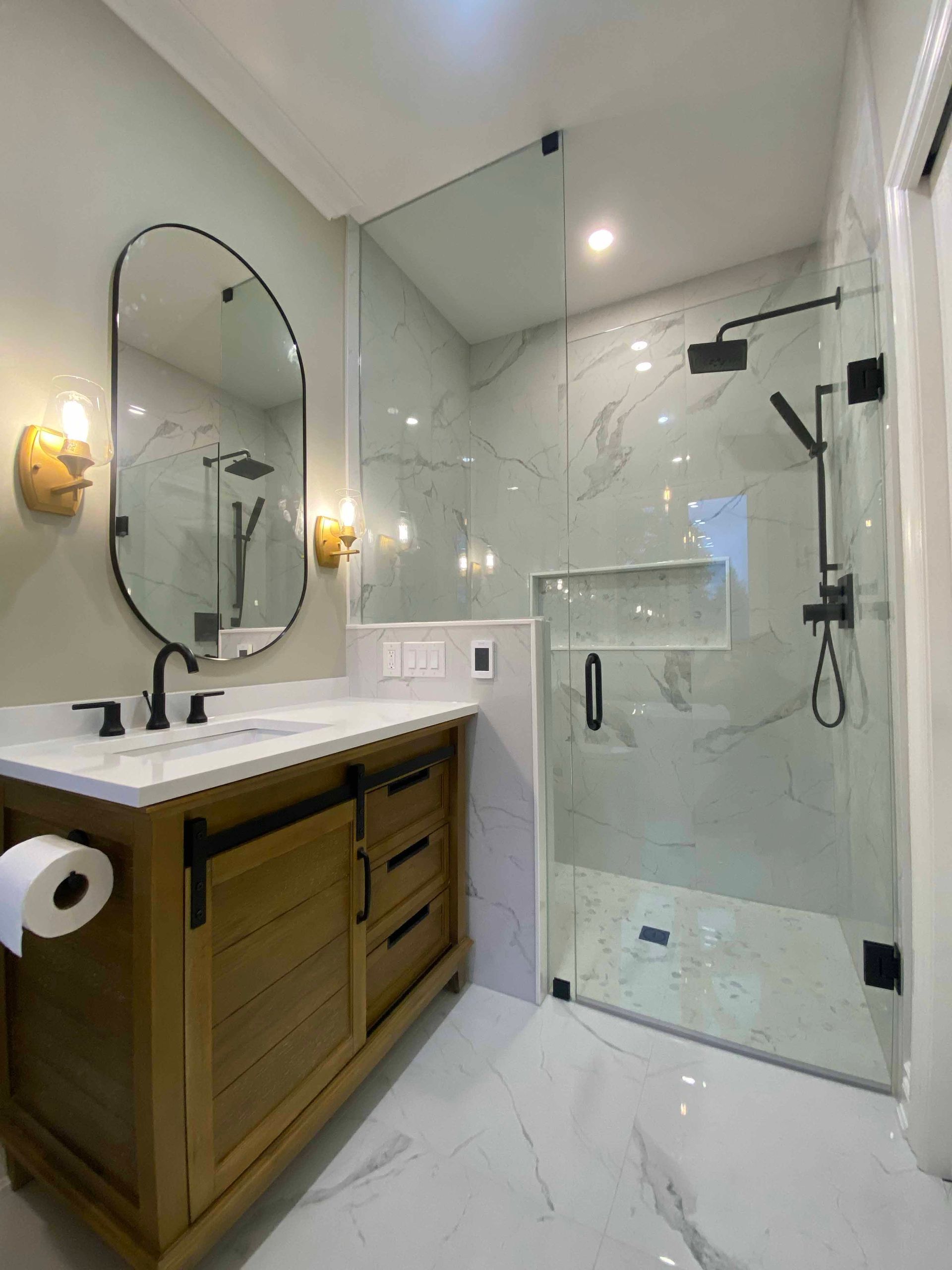 Modern bathroom with wooden vanity, oval mirror, and glass walk-in shower with white marble-style tile.