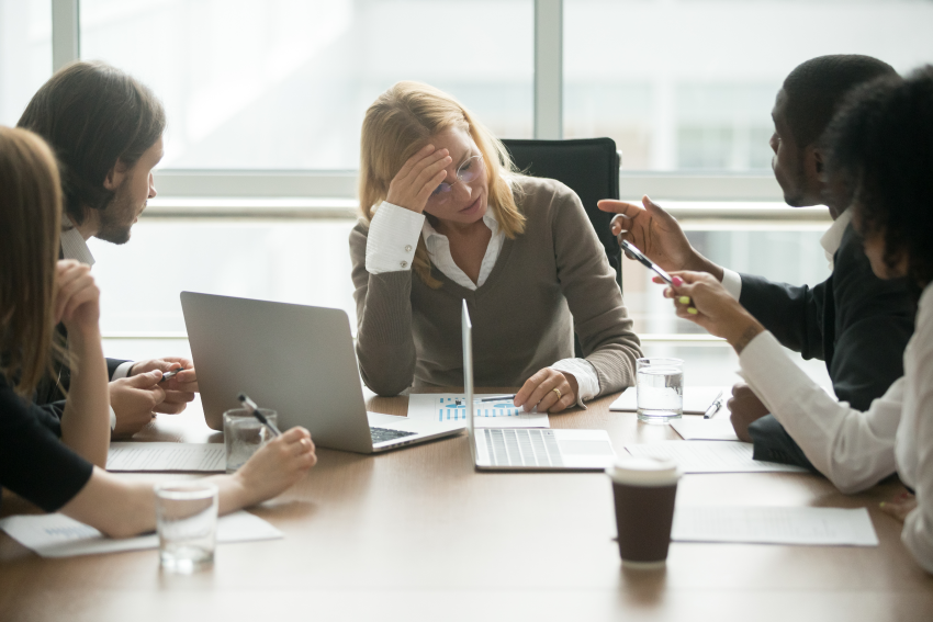 Woman looking stressed at a table with coworkers in an office setting, some pointing and gesturing.
