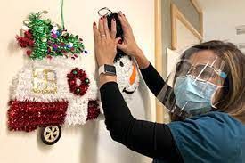 Person in teal scrubs and face shield decorates a wall with Christmas ornaments.