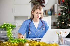 Nurse in blue scrubs, stethoscope, working on a laptop at a desk decorated for Christmas.