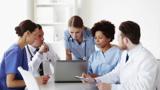 a group of doctors and nurses are sitting around a table looking at a laptop .