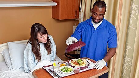 A man is serving food to a woman in a hospital bed.