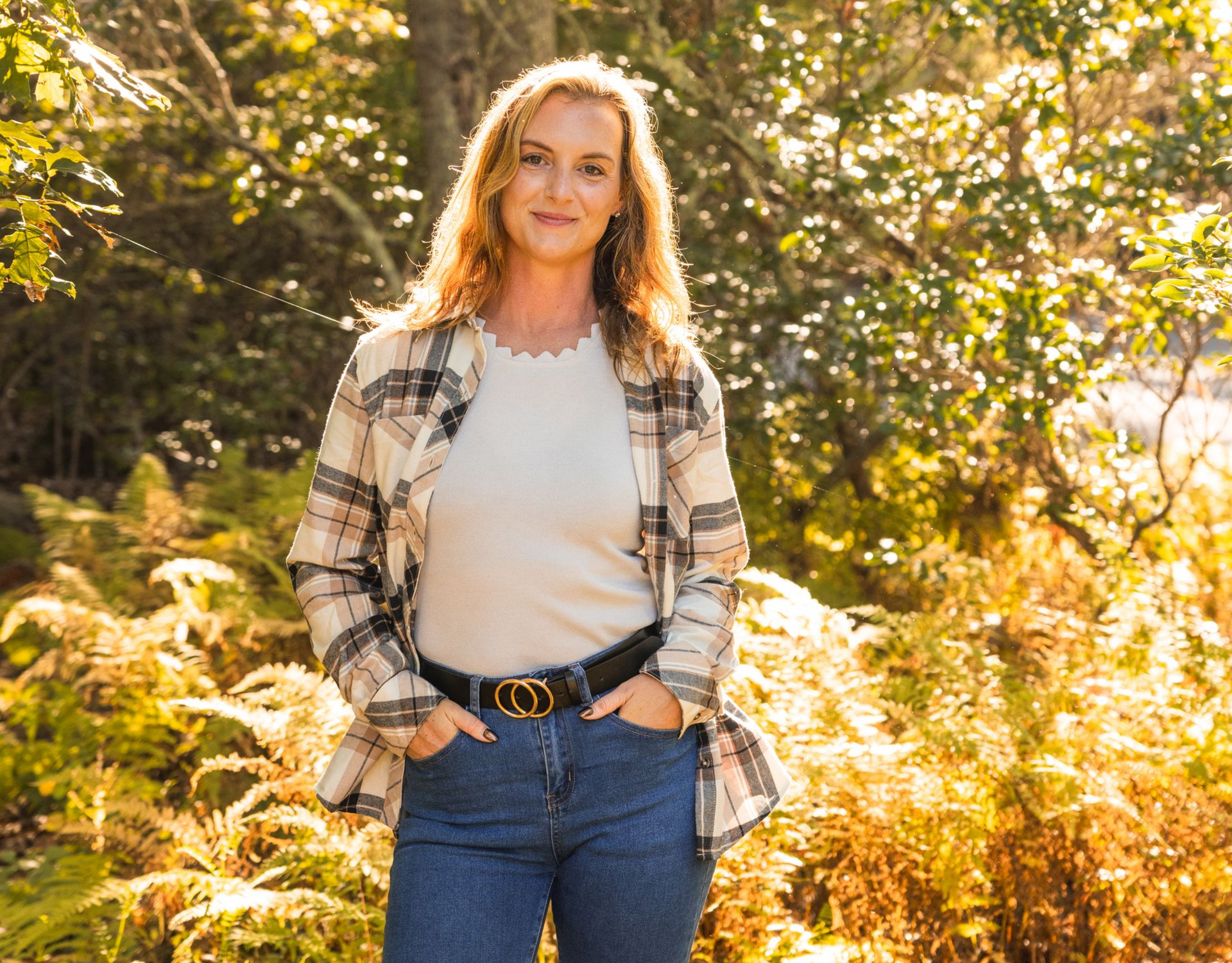 Person standing outdoors in a sunlit field, wearing a plaid shirt, white top, and jeans, with hands in pockets.