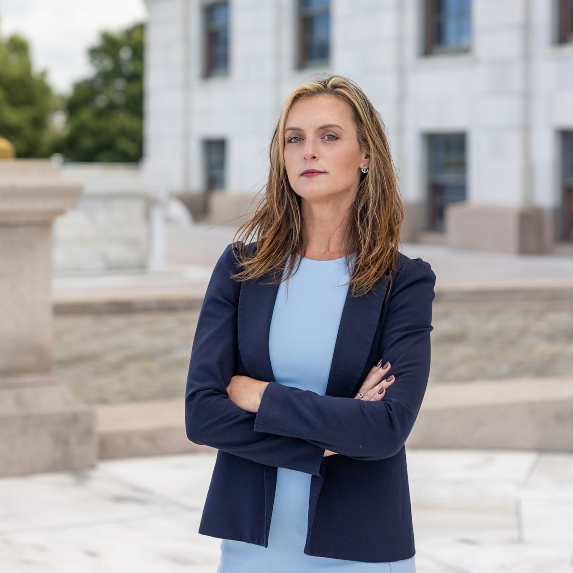 Woman in navy blazer and light blue dress standing with arms crossed outside a modern building