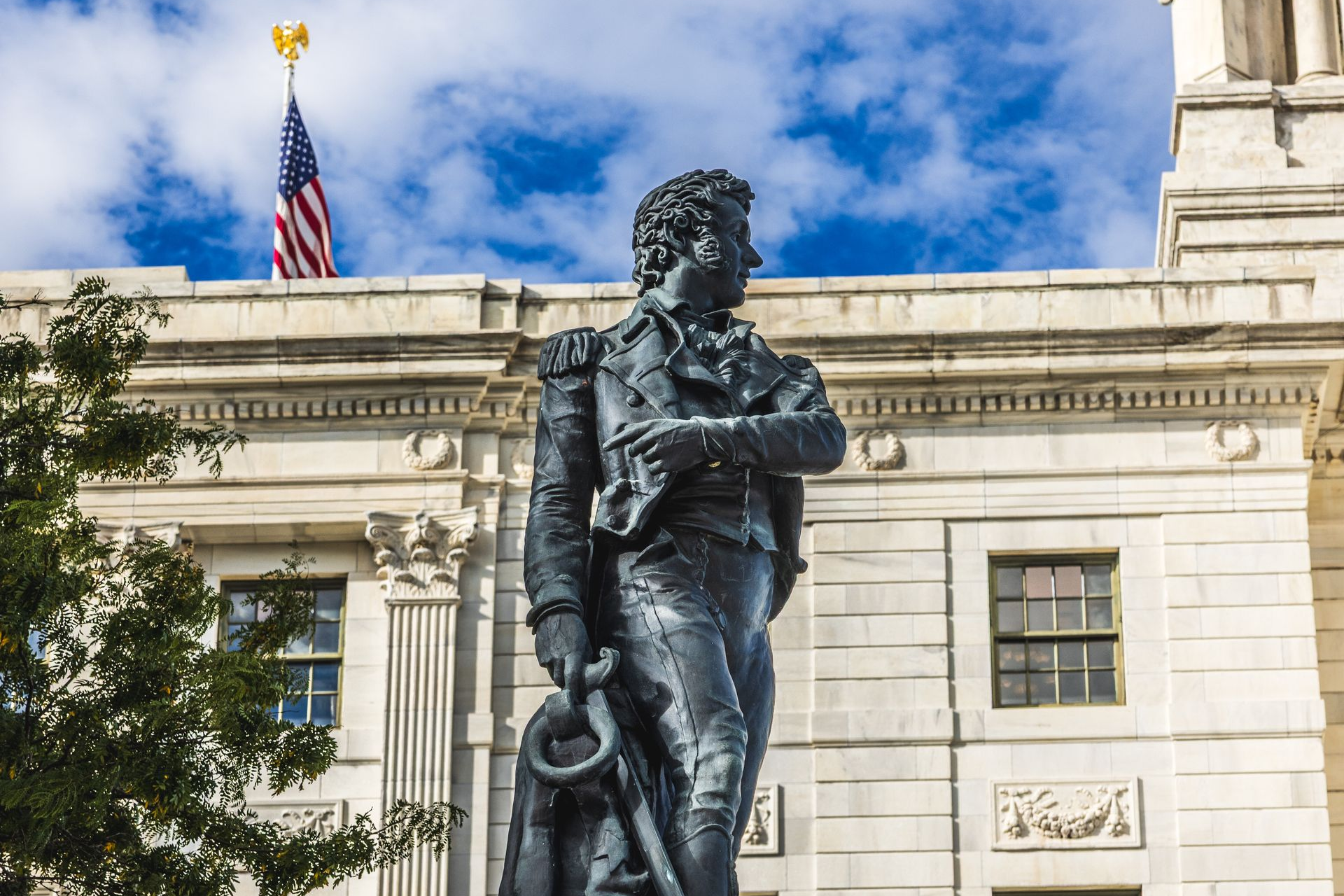 Bronze statue before a white stone building with an American flag under a blue sky